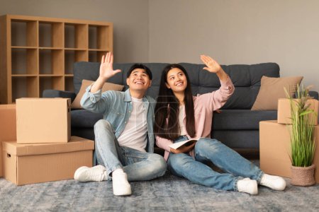 Photo for House Repairment. Cheerful Asian Couple Planning Renovation In New Apartment After Moving In, Sitting On Floor Among Carton Boxes And Framing Room Interior With Hands At Modern Living Indoor - Royalty Free Image