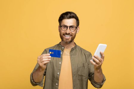 Téléchargez les photos : Banque virtuelle, e-commerce. Heureux homme d'âge moyen européen avec smartphone et carte de crédit bancaire dans ses mains posant sur fond jaune, souriant à la caméra - en image libre de droit