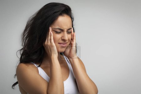 Verstörte unglückliche Frau aus dem Mittleren Osten, die Schmerzen an den Schläfen hat, während der Periode unter Kopfschmerzen Migräne leidet, isoliert auf grauem Hintergrund, Nahaufnahme, Kopierraum