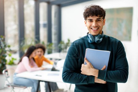 Photo for Happy european student guy posing with notepads in hands and headphones on neck, smiling at camera while female classmates studying in coworking space interior - Royalty Free Image