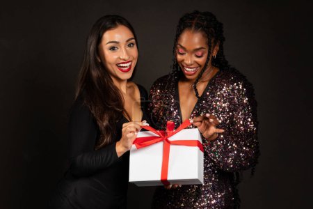Téléchargez les photos : Cadeau de Noël. Joyeux multiethnique belles jeunes femmes copines tenant boîte cadeau posant sur fond de studio noir. Célébration des fêtes d'hiver, célébration du Nouvel An et concept de cadeaux de Noël - en image libre de droit