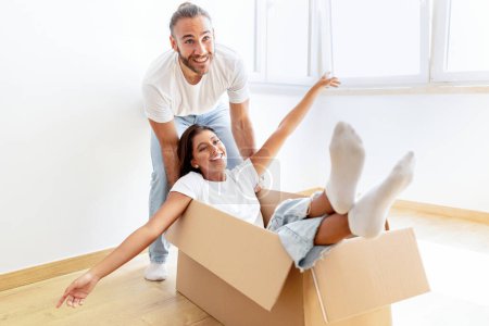 Photo for Playful young multiracial couple having fun in their home on relocation day. Millennial woman and her boyfriend playing together with carton box while moving to new apartment, changing house - Royalty Free Image