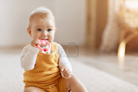 Photo for Closeup of adorable little baby girl sitting on floor warm soft carpet at home, chewing pink teether and looking at copy space. Children Oral-Motor Sensory Chews, Kid Toys for Chewing - Royalty Free Image