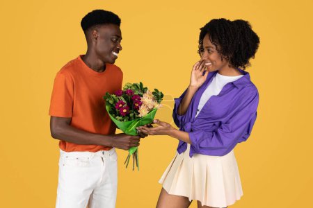 Photo for Positive millennial black guy gives bouquet of flowers to lady, isolated on yellow studio background. Birthday greeting, anniversary surprise, holiday celebration together and romantic date - Royalty Free Image
