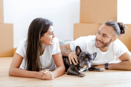 Photo for Happy excited beautiful young multicultural couple and their dog french bulldog moving to new house, millennial man and woman lying on floor in empty apartment, unpacking boxes, caressing puppy - Royalty Free Image