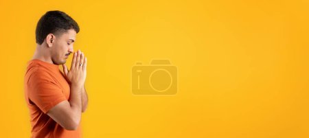 Téléchargez les photos : Vue latérale de l'homme brésilien priant tenant les paumes par les lèvres et chuchotant priant, demandant grâce sur fond de studio jaune, panorama avec espace de copie - en image libre de droit