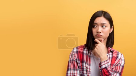 Photo for Indecisive pensive asian young lady posing on yellow studio background, touching chin and looking aside at copy space for advertisement, panorama, banner - Royalty Free Image