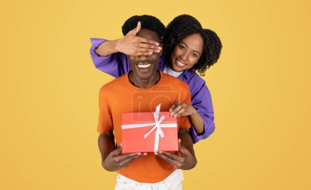 Photo for Positive millennial black lady closes eyes to boyfriend with gift box, isolated on yellow studio background. Birthday greeting, anniversary surprise, holiday celebration together - Royalty Free Image