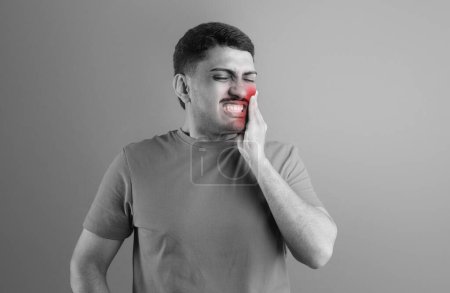 Téléchargez les photos : Problème de dents. Jeune homme souffrant de maux de dents aigus, touchant la zone douloureuse rouge, frottant la mâchoire, parodontite, debout sur le fond du studio, prise de vue BW - en image libre de droit