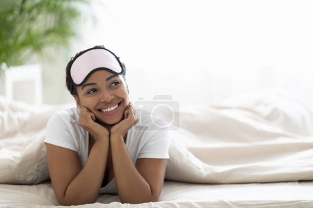 Téléchargez les photos : Heureuse jeune femme noire portant un masque de sommeil sur la tête en regardant l'espace de copie tout en se relaxant sur le lit à la maison, détendue belle afro-américaine se reposant dans la chambre confortable intérieur après le réveil - en image libre de droit
