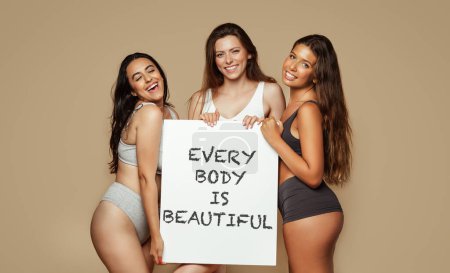 Photo for Positive young multiethnic women in different underwear, enjoy natural beauty, hold banner inscription every body is beautiful, isolated on beige background, studio. Lifestyle, health care, love - Royalty Free Image