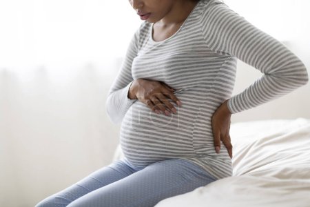 Photo for Pregnancy Pain. Cropped Shot Of Black Pregnant Lady Feeling Unwell At Home, African American Female Sitting On Bed, Touching Belly And Lower Back, Suffering Prenatal Contractions, Free Space - Royalty Free Image