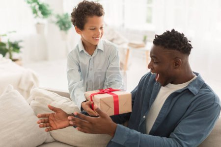 Foto de Niño sonriente sosteniendo caja de regalo saludando papá afroamericano, felicitándolo en el cumpleaños, celebrando las vacaciones familiares con regalos en el interior del hogar moderno. Celebración del día de los padres - Imagen libre de derechos