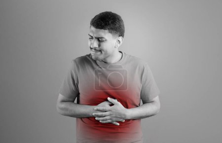 Foto de Hombre brasileño enfermo que tiene dolor de estómago, tocándose el abdomen con la zona inflamada roja, que sufre de enfermedad gastrointestinal, posando en el fondo del estudio, foto en blanco y negro - Imagen libre de derechos