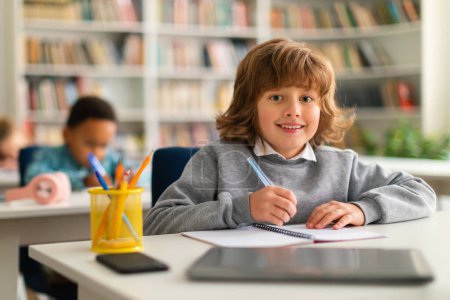 Photo for Positive smart schoolboy sitting at desk in classroom, smiling at camera while writing in notebook, classmates studying on the background - Royalty Free Image