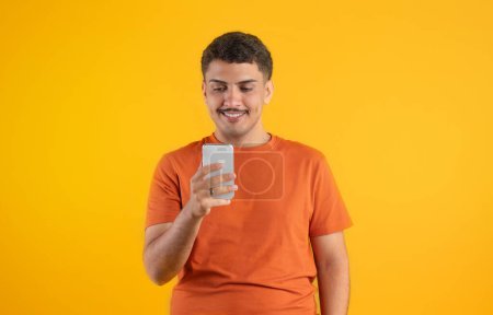 Photo for Positive brazilian man using cellphone and browsing internet, scrolling social media news feed or texting online over yellow background. People and gadgets - Royalty Free Image