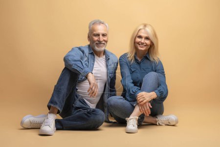 Photo for Happy senior man and woman couple friends wearing denim outfit sitting on floor over beige background, smiling at camera. Cheerful elderly spouses posing together, studio shot. Life after retirement - Royalty Free Image