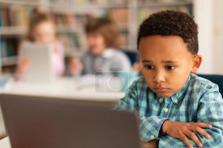Téléchargez les photos : Ennuyé grave enfant noir faire des devoirs ou regarder la vidéo sur ordinateur portable, assis au bureau en classe avec des camarades de classe sur fond flou - en image libre de droit
