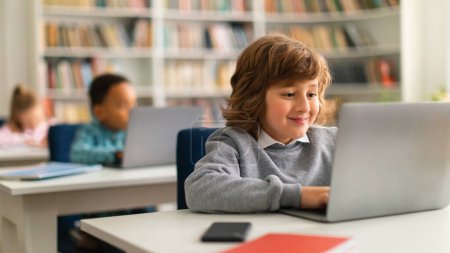 Téléchargez les photos : Un écolier européen intelligent souriant assis au bureau devant un ordinateur portable en classe, tapant ou faisant ses devoirs en ligne, panorama. Enseignement primaire moderne - en image libre de droit