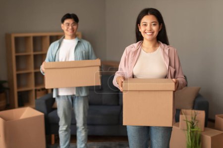 Photo for Real Estate Offer. Young Asian Spouses Holding Cardboard Boxes Happy About Moving, Posing Looking at Camera and Smiling in New Home. Millennial Couple Settling in Rented Apartment. Selective Focus - Royalty Free Image