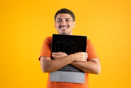 Foto de Hombre brasileño excitado en casual abrazando portátil moderno PC con pantalla en blanco negro y sonriendo a la cámara, posando sobre fondo de estudio amarillo. Amor dispositivo agradable gadget - Imagen libre de derechos