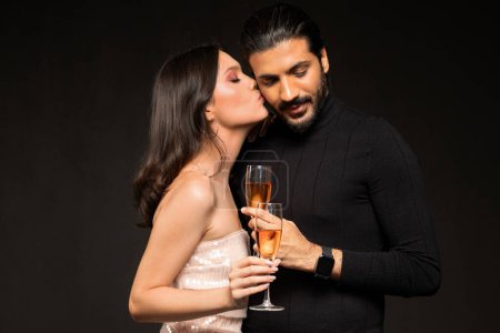 Photo for Loving multicultural young couple wearing beautiful outfits drinking champagne on black background, pretty brunette woman kissing indian guy boyfriend, lovers celebrating New Year, Christmas - Royalty Free Image