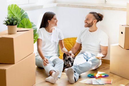 Photo for Relocation. Excited multicultural young couple sitting on floor among cardboxes with belongings in empty house, stroking their pet dog, have conversation, discussing interior design for new apartment - Royalty Free Image