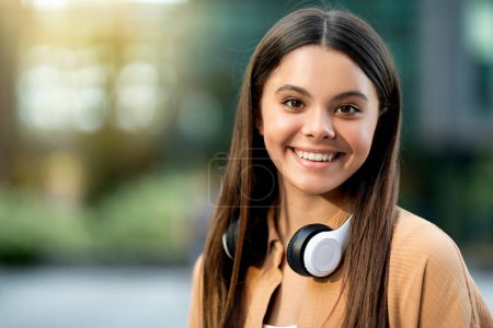 Téléchargez les photos : Portrait d'une jolie jeune femme hispanique brune joyeuse portant une tenue décontractée et un casque sans fil étudiant posant sur le campus de l'université, souriant à la caméra. Éducation, école, concept universitaire - en image libre de droit
