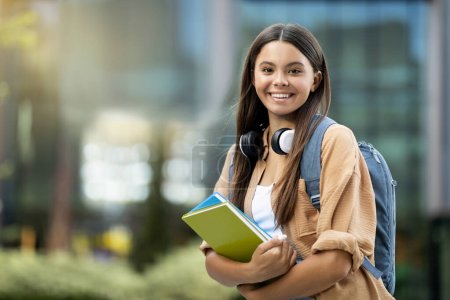 Photo for Happy beautiful long-haired hispanic young lady in casual outfit with wireless headphones and backpck going to college, posing at university campus, holding exercise books, copy space - Royalty Free Image