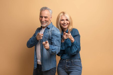 Photo for Confident cool attractive caucasian elderly man and woman wearing casual denim outfit gesturing pointing at camera and smiling, isolated on beige background. Successful retirees - Royalty Free Image