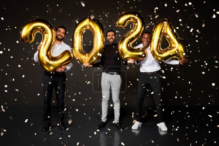 Foto de Felices chicos jóvenes multirraciales vistiendo bonitos trajes celebrando el año nuevo 2024 juntos sobre fondo negro, diversos millennials hombres sosteniendo globos dorados y sonriendo a la cámara - Imagen libre de derechos