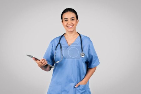 Téléchargez les photos : Portrait de médecin gai dame latine en uniforme bleu tenant presse-papiers et souriant à la caméra, posant sur fond gris. Offre médicale publicité, soins de santé - en image libre de droit
