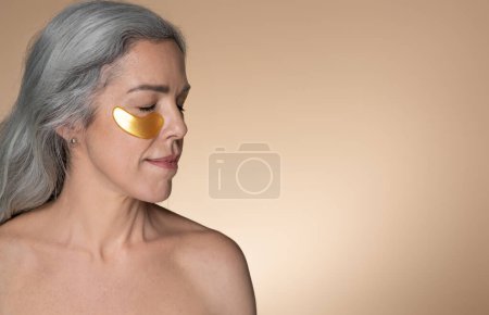 Téléchargez les photos : Femme blanche séduisante aux cheveux gris bénéficiant d'un traitement anti-âge, appliquant des patchs oculaires, posant sur fond beige. Produits cosmétiques hydratants, soins de beauté et spa - en image libre de droit