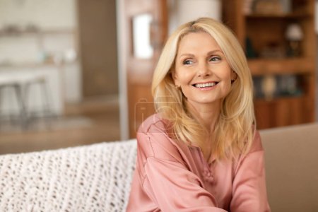 Téléchargez les photos : Portrait de belle blonde mature dame souriante regardant à l'extérieur posant sur le canapé à la maison. Gros plan de attrayant femme jouissant de sa beauté et le vieillissement dans confortable intérieur domestique, Copier l'espace - en image libre de droit
