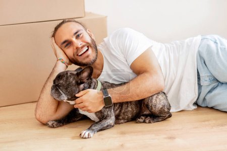 Photo for Happy stylish millennial guy hipster lying on floor in empty house among cardboxes with his belongings, caressing his pet dog in new apartment, enjoying new house. Rent, relocation, moving - Royalty Free Image