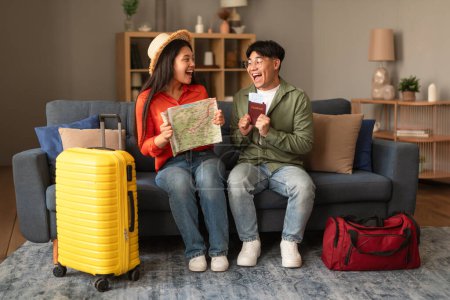 Foto de Alegría de vacaciones. Emocionados turistas coreanos Pareja sosteniendo mapa de viajes y entradas, sentado con la maleta, celebrando el viaje a un nuevo destino en casa, sonriendo el uno al otro. Viajeros listos para el vuelo - Imagen libre de derechos