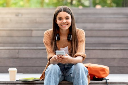 Photo for Portrait of cheerful beautiful hispanic young woman student using phone outtdoor, sitting on staits at city urban area, drinking takeaway coffee, getting ready for class, waiting for friends - Royalty Free Image