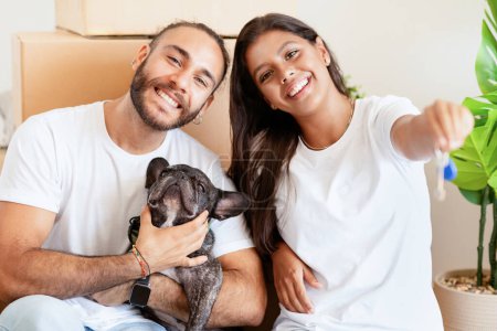 Photo for Cheerful young multiethnic couple with cute french bulldog dog sitting on floor in new home on relocation day. Happy millennial husband and wife with cute pet moving to new house, showing key - Royalty Free Image