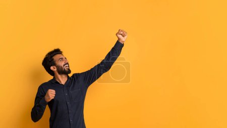 Téléchargez les photos : Hourra Concept. Joyeux jeune Indien célébrant le succès avec le poing levé, heureux Euphorique oriental Guy acclamant Gagner, heureux mâle debout sur fond jaune Studio, Panorama avec espace de copie - en image libre de droit