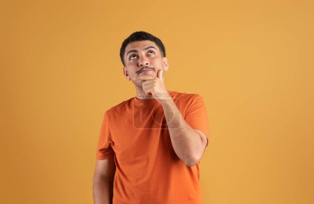 Téléchargez les photos : Curieux homme brésilien coûteux touchant menton regardant vers le haut à l'espace de copie pour la publicité, posant sur fond de studio orange, offre intéressante - en image libre de droit