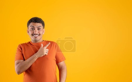 Téléchargez les photos : Cool joyeux sourire bel homme brésilien en t-shirt pointant de côté à l'espace de copie pour la publicité et souriant à la caméra sur fond jaune studio. Bannière Web pour l'offre en ligne - en image libre de droit
