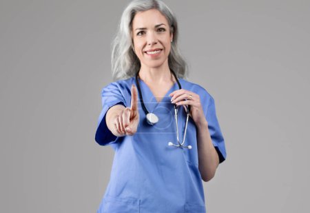 Téléchargez les photos : Femme infirmière d'âge moyen souriante en tenue de travail bleue avec stéthoscope pointant du doigt vers le haut, ayant des conseils médicaux pour vous sur fond de studio gris. Soins de santé, concept de médecine. Concentration sélective - en image libre de droit