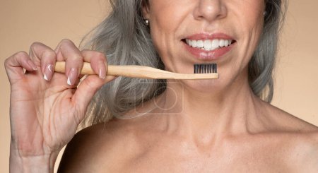 Foto de Mujer mayor feliz utilizando el cepillo de dientes de bambú natural ecológico para limpiar los dientes, disfrutando de artículos de plástico gratis para la higiene personal, de pie sobre fondo beige, primer plano - Imagen libre de derechos
