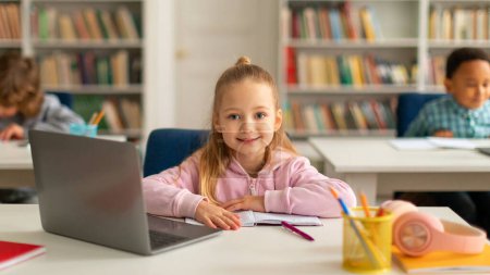 Photo for Happy caucasian school girl using laptop computer at class, looking and smiling at camera, school student learning online using technology during tech lesson - Royalty Free Image