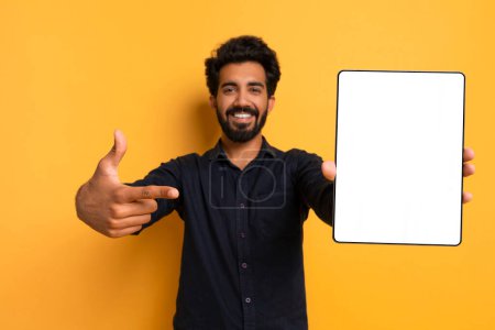Foto de Oferta en línea. Hombre indio guapo apuntando a la tableta digital con pantalla blanca, sonriendo joven oriental que demuestra espacio libre de la copia para el diseño del anuncio, de pie sobre fondo amarillo, burla - Imagen libre de derechos