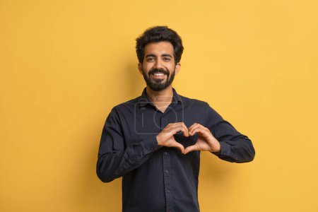 Photo for Smiling Young Indian Man Showing Heart Gesture With Hands Near Chest, Happy Millennial Eastern Guy In Black Shirt Making Love Sign At Camera While Posing Over Yellow Studio Background, Copy Space - Royalty Free Image