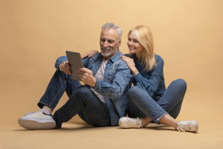 Téléchargez les photos : Couple sénior positif assis sur le sol, embrassant et utilisant une tablette numérique PC moderne, isolé sur fond de studio beige. Homme et femme retraités wesurf, scrolling, shopping en ligne sur pad - en image libre de droit