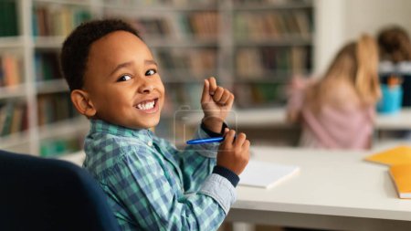 Photo for Positive black primary schoolboy sitting at desk in classroom, looking and smiling at camera, panorama, free space. Classmates studying on the background - Royalty Free Image