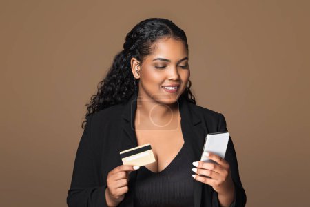 Photo for Financial offer. Cheerful brazilian plus size woman using cellphone and credit card, advertising bank service over brown studio background. Female customer posing with phone and bankcard - Royalty Free Image