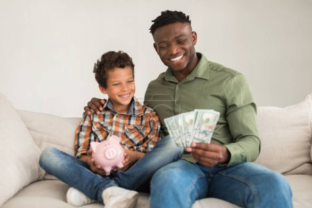 Foto de Feliz padre negro y su hijito posando con dinero en efectivo y dinero, sosteniendo billetes de dólar que anuncian una gran oferta bancaria, sentado en el sofá en casa. Concepto de Inversiones Financieras - Imagen libre de derechos